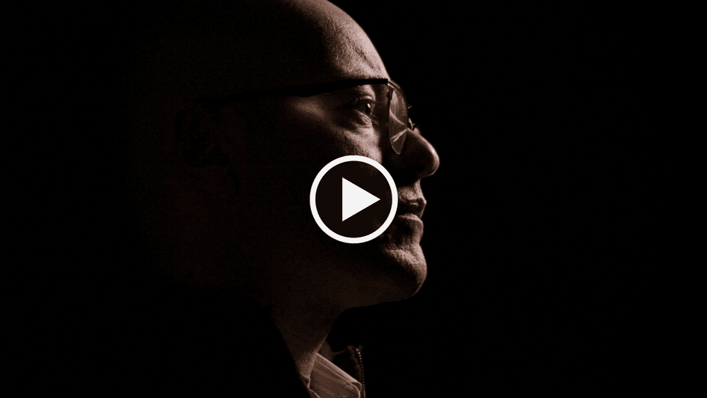 Dark side profile of a bald man with glasses, with a white play button over the image